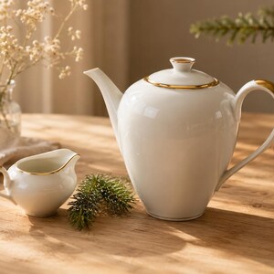 May include: A white porcelain teapot and creamer set with gold detailing, presented on a wooden table. The teapot has a rounded form with a lid and a curved handle. The creamer is smaller, with a matching design. A lit candle and greenery are in the background.
