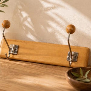 Könnte beinhalten: Garderobenleiste aus Holz mit zwei Metallhaken und hölzernen Kugelfüßen. Die Leiste ist auf einer hellen Holzplatte montiert. Eine Vase mit Grün, eine kleine Holzschale und ein geflochtener Korb sind ebenfalls zu sehen.
