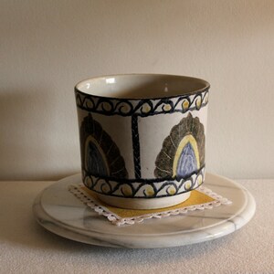 May include: A ceramic cup with a white base and a decorative pattern of dark green, blue, and yellow floral designs. The cup sits on a marble coaster with a yellow square doily. The cup is approximately 4 inches tall.