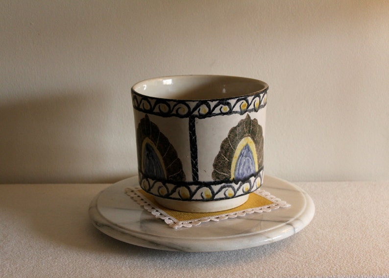 May include: A ceramic cup with a white base and a decorative pattern of dark green, blue, and yellow floral designs. The cup sits on a marble coaster with a yellow square doily. The cup is approximately 4 inches tall.