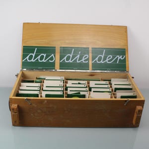 Puede incluir: Una caja de madera vintage con tapa con bisagras, que contiene numerosas tarjetas pequeñas. La tapa presenta tres pizarras verdes con las palabras "das die der" escritas en cursiva blanca. La caja está abierta, revelando las tarjetas organizadas.