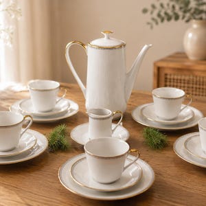 Puede incluir: Un juego de té de porcelana blanca con ribete dorado, que incluye una tetera, una jarra de crema, un azucarero y seis tazas con platillos. El juego está dispuesto sobre una mesa de madera, con un fondo neutro y luz natural.