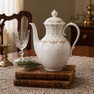 May include: A white porcelain teapot with a floral design and lid rests on old books. The teapot is part of a tea set, with plates on the table. A vase of flowers, a candle, and a wine glass are also on the table, creating a classic tea setting.