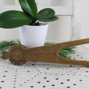May include: A vintage brass nutcracker with intricate carvings and a floral design. The nutcracker is lying on a white surface with a potted plant in the background.