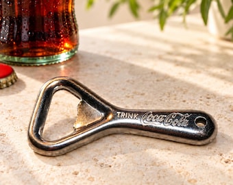 May include: A metallic bottle opener featuring the embossed text TRINK Coca-Cola on its handle. The tool has a triangular head for prying off bottle caps and a small circular hole at the end of the handle for hanging or storage purposes.