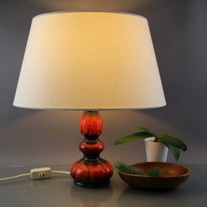 May include: A ceramic table lamp with a cream lampshade and a red and black base. The lamp has a bulbous shape and a textured surface. A small wooden bowl and a potted plant are next to the lamp.