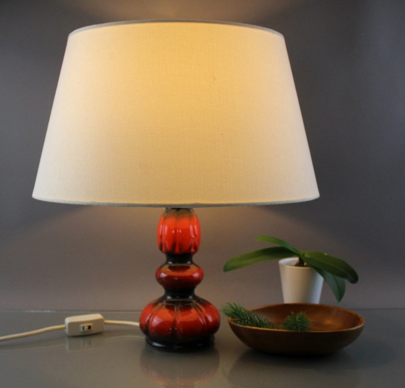 May include: A ceramic table lamp with a cream lampshade and a red and black base. The lamp has a bulbous shape and a textured surface. A small wooden bowl and a potted plant are next to the lamp.