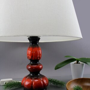 May include: A ceramic table lamp with a white lampshade. The lamp base is a glossy red and black colour. The lamp is on a reflective surface with a small plant and wooden bowl in the background.
