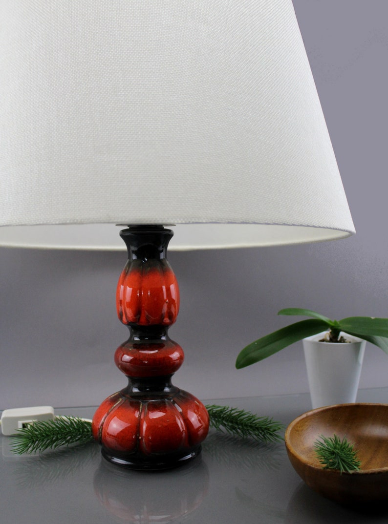 May include: A ceramic table lamp with a white lampshade. The lamp base is a glossy red and black colour. The lamp is on a reflective surface with a small plant and wooden bowl in the background.