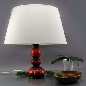 May include: A retro table lamp featuring a white lampshade and a red and black ceramic base. The lamp is illuminated, with the power cord visible. A small wooden bowl and a potted plant are also in the image.