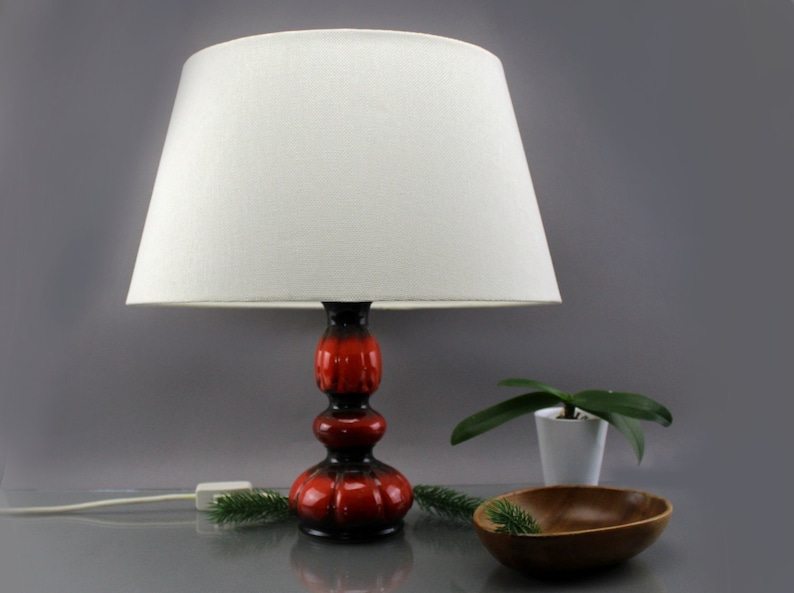 May include: A retro table lamp featuring a white lampshade and a red and black ceramic base. The lamp is illuminated, with the power cord visible. A small wooden bowl and a potted plant are also in the image.