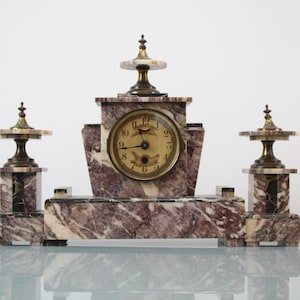 Puede incluir: Reloj antiguo de mármol con una esfera central y dos elementos decorativos laterales. El reloj presenta un patrón de mármol marrón y blanco con detalles en latón. La esfera del reloj tiene números y agujas negras.