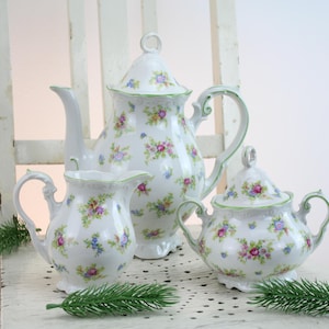Puede incluir: Juego de té de porcelana blanca con tetera, jarra de crema y azucarero. El juego está decorado con un estampado floral en rosa, morado y azul, con ribetes verdes. La tetera mide unos 20 cm de alto.
