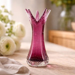 Könnte beinhalten: Eine elegante, magenta-farbene Glasvase mit sternförmiger Spitze. Die Vase hat einen schlanken Hals und einen abgerundeten Boden mit Luftblasen. Im Hintergrund weiße Blumen und eine Holzoberfläche. Die Vase ist etwa 20 cm hoch.