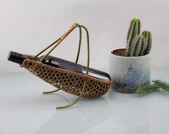 Puede incluir: Un porta botellas de vino de metal dorado con una cesta de mimbre tejida. El soporte tiene un diseño curvo y tres patas. Una botella de vino descansa en la cesta.