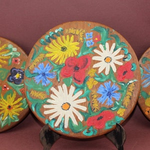 May include: Three round wooden plates with hand-painted floral designs. The plates feature a variety of flowers in shades of red, yellow, blue, white, and green.
