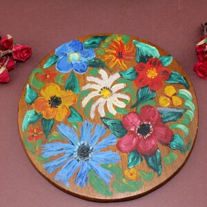 May include: A round wooden coaster with a painted floral design. The coaster features a variety of flowers in shades of red, yellow, blue, and white.