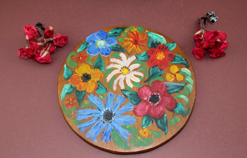 May include: A round wooden coaster with a painted floral design. The coaster features a variety of flowers in shades of red, yellow, blue, and white.