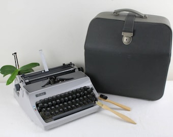 Op de afbeelding: Een zilveren typemachine met een zwart toetsenbord en een zwarte draagtas. De typemachine heeft een plant erop en twee houten borstels ervoor.