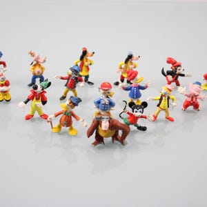 May include: A collection of colorful miniature figurines of cartoon characters, including Mickey Mouse, Donald Duck, and Goofy. The figures are in various poses and outfits, with bright colors and detailed features. The figurines are arranged on a light gray surface.