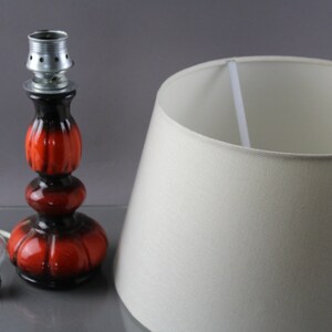 May include: A retro table lamp featuring a red and black ceramic base and a cream lampshade. The lamp includes a light bulb and a power cord. The base has a bulb socket. The lampshade is a classic conical shape.