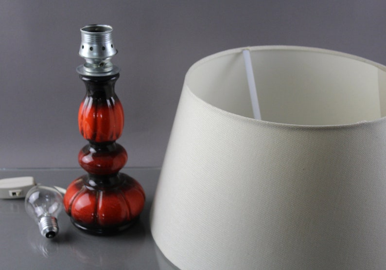 May include: A retro table lamp featuring a red and black ceramic base and a cream lampshade. The lamp includes a light bulb and a power cord. The base has a bulb socket. The lampshade is a classic conical shape.