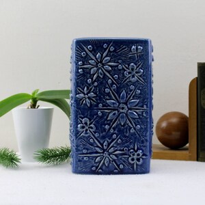 May include: A blue ceramic vase with a raised floral design. The vase is rectangular and has a textured surface.