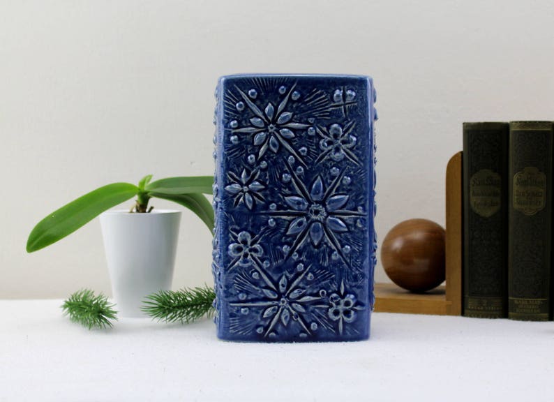 May include: A blue ceramic vase with a raised floral design. The vase is rectangular and has a textured surface.