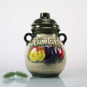 May include: A vintage ceramic Rumtopf jar with a dark green lid and handles. The jar is decorated with colourful fruit illustrations and the word "Rumtopf" in script. The jar has a textured, dotted pattern on the body.