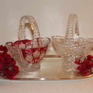 Puede incluir: Dos cestas de cristal con asas, una con un diseño rojo y blanco, la otra transparente, sobre una bandeja plateada con flores de seda rojas.