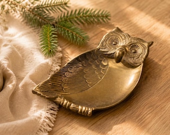 May include: A brass-colored metal trinket dish shaped like an owl. The dish features detailed engravings of feathers on the wings and a textured face with prominent eyes and a beak. The surface has a metallic finish with dark antique-style highlights.