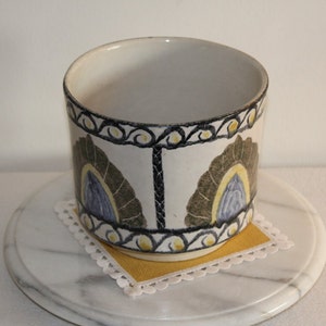 May include: A cylindrical ceramic container with a white base, adorned with a repeating pattern of stylized feather-like shapes in olive green, blue, and yellow. The rim features a black and yellow scroll design. The container sits on a yellow square doily.