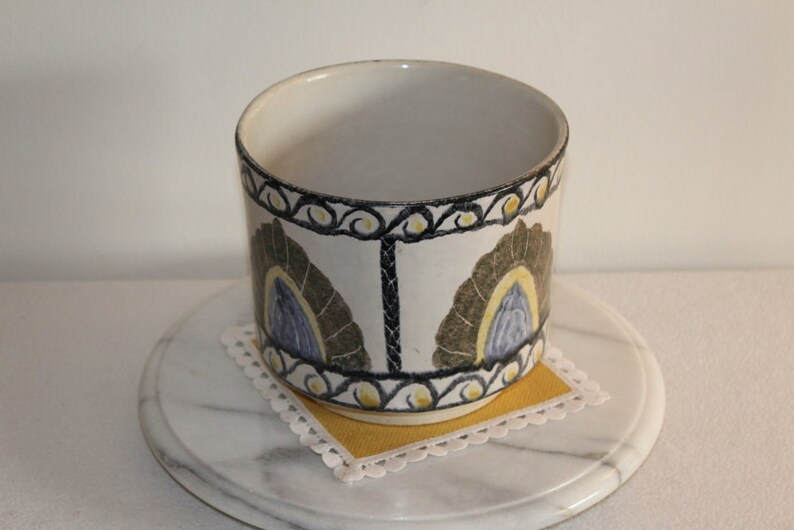 May include: A cylindrical ceramic container with a white base, adorned with a repeating pattern of stylized feather-like shapes in olive green, blue, and yellow. The rim features a black and yellow scroll design. The container sits on a yellow square doily.