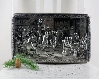 May include: A silver metal box with a raised design of people dancing in a room. The box has a decorative border around the edge.