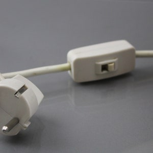 May include: A close-up shot of a white electrical plug with three pins and a white cable. A white inline switch is attached to the cable. The items are on a grey surface.
