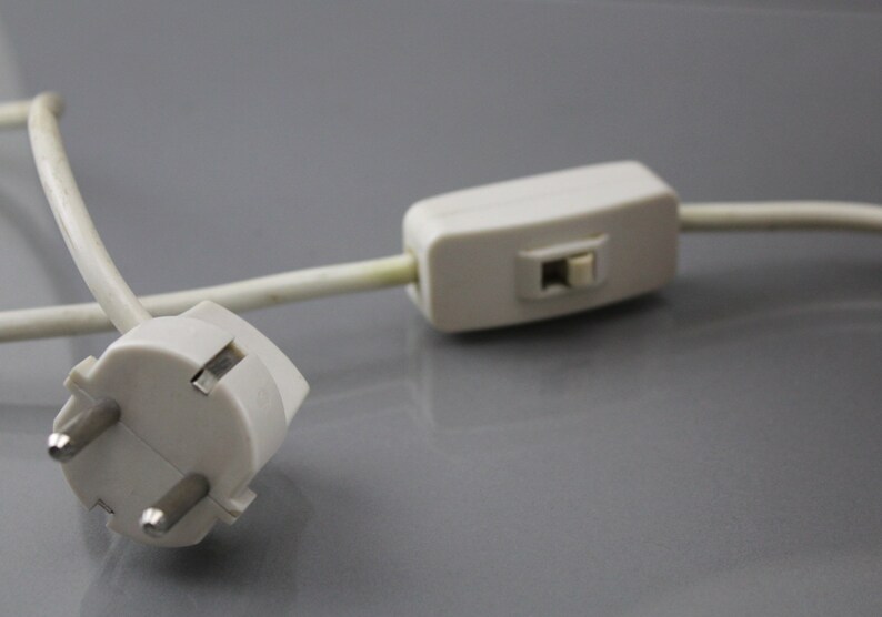 May include: A close-up shot of a white electrical plug with three pins and a white cable. A white inline switch is attached to the cable. The items are on a grey surface.
