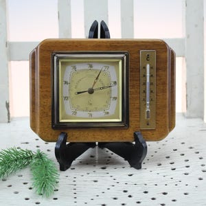 May include: A vintage wooden weather station with a clock, barometer, and thermometer. The clock face is cream-coloured with black hands and numerals. The thermometer is in Celsius. The unit is mounted on a black stand.
