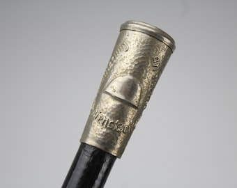 May include: A walking stick handle made of silver-toned metal with a hammered texture. The handle features a raised semi-circular emblem and engraved German text that reads "Dienstzeit" and "Erinnerung an meine". The shaft is black.