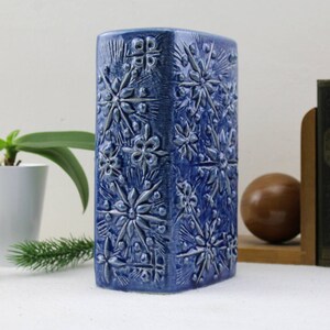 May include: A blue ceramic vase with a raised floral pattern. The vase is rectangular and has a textured surface.