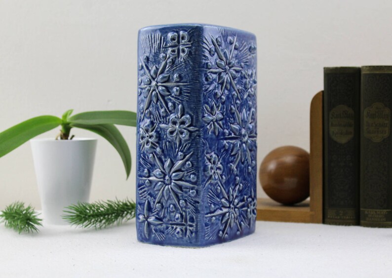 May include: A blue ceramic vase with a raised floral pattern. The vase is rectangular and has a textured surface.