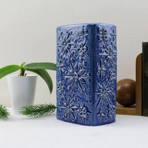 May include: A blue ceramic vase with a geometric pattern of stars. The vase is rectangular and has a smooth, glazed finish.
