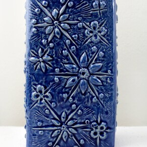 May include: A blue ceramic vase with a textured surface featuring a repeating pattern of embossed flowers and stars.