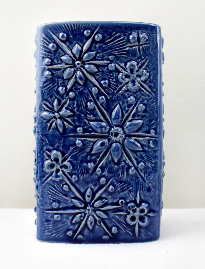 May include: A blue ceramic vase with a textured surface featuring a repeating pattern of embossed flowers and stars.