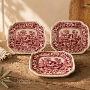 May include: Three square plates with a red and white landscape pattern and a scalloped edge. They are arranged on a wooden surface. A vase with greenery and a linen cloth are also visible. The plates are likely ceramic.