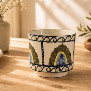 May include: A ceramic cup with a cream-colored base and a decorative pattern. The design includes blue, yellow, and green fan-shaped motifs, and a dark blue border. The cup is on a wooden table, with a vase of flowers and a wooden bowl in the background.