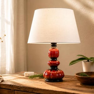 May include: A table lamp featuring a red and black ceramic base and a white lampshade. The lamp sits on a wooden table, alongside a small plant, a wooden bowl, and a vase with dried flowers. The lamp is a home decor item.