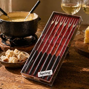 May include: A fondue set with a black pot, forks, and a box of fondue forks. The set includes a bowl of bread cubes, cheese, and wine glasses. The image is set on a wooden table.