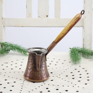 Könnte beinhalten: Eine kleine Kaffeekanne aus Kupfer mit gehämmerter Oberfläche und einem Holzgriff. Die Kanne steht auf einer weißen Oberfläche mit einem gemusterten Design.