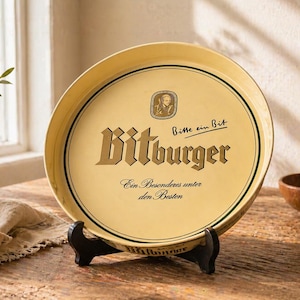 Könnte beinhalten: Ein runder, gelber Bitburger Servierteller auf einem schwarzen Ständer. Der Teller zeigt das Bitburger-Logo in goldfarbenen Buchstaben und deutschen Text. Ein Holzschneidebrett, eine kleine Schüssel und eine Vase mit Grünzeug befinden sich im Hintergrund.