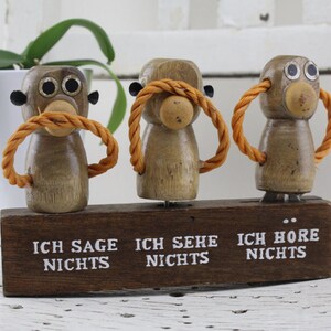 May include: Three wooden monkeys with orange rope loops around their mouths and large black eyes. The monkeys are standing on a wooden base with the text "Ich sage nichts", "Ich sehe nichts", and "Ich höre nichts" written on it.